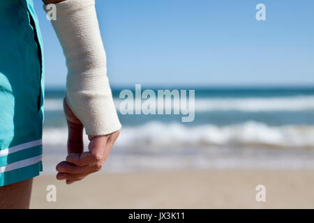 Primo piano di un giovane uomo caucasico in costume da bagno sulla spiaggia con un bendaggio nel suo polso, di fronte al mare con lo spazio negativo sul lato destro Foto Stock