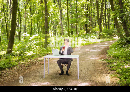 Imprenditore seduto alla scrivania in ufficio con computer portatile e la tazza di caffè parlando cellulare nella foresta verde parco. Foto Stock