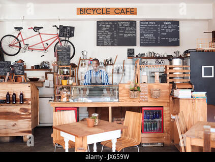 Sorridente imprenditore africani in piedi dietro un contatore del suo cafe Foto Stock