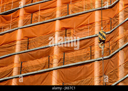 I cavi della gru e il gancio contro la maglia arancione di un edificio in costruzione dello sfondo. Foto Stock