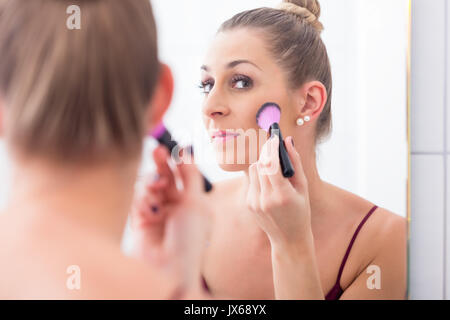 Donna spolvero il suo volto con spazzola di cosmetici di fronte a Specchio bagno Foto Stock