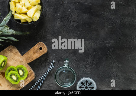 Kiwi freschi fette su tavola di legno e ananas tagliato a pezzi in ciotola su sfondo scuro. Vista superiore, copia dello spazio. Preparazione frullato Foto Stock