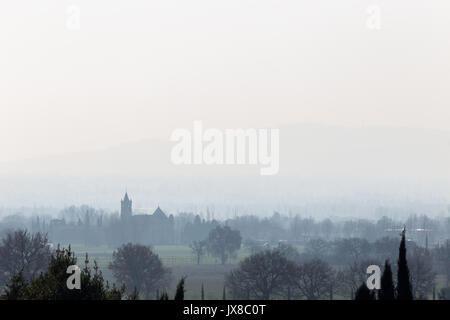 Una vista della città di Rivotorto (vicino ad Assisi, Umbria, Italia) nel mezzo della nebbia, con silhouette della chiesa e molti alberi intorno Foto Stock