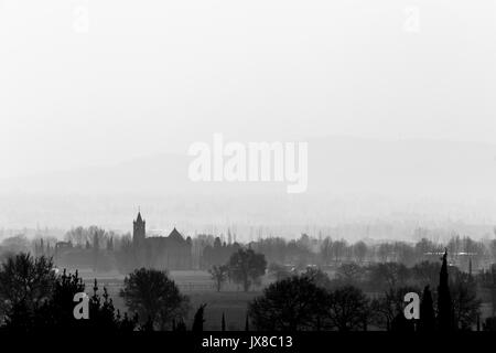 Una vista della città di Rivotorto (vicino ad Assisi, Umbria, Italia) nel mezzo della nebbia, con silhouette della chiesa e molti alberi intorno Foto Stock