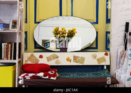 Spazio alla creatività. Casa accogliente interno da uno specchio ovale, libri, fiori gialli di garofani in un vaso e un divano con cuscini contro lo sfondo di una porta di colore giallo con strisce blu. Foto Stock