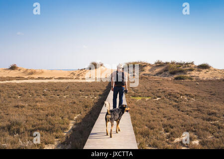 Vista da dietro di un uomo che cammina con il suo cane su una strada che conduce attraverso il bellissimo paesaggio. Foto Stock