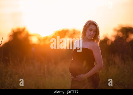 Donna incinta in un abito nero con un tramonto dietro di lei Foto Stock