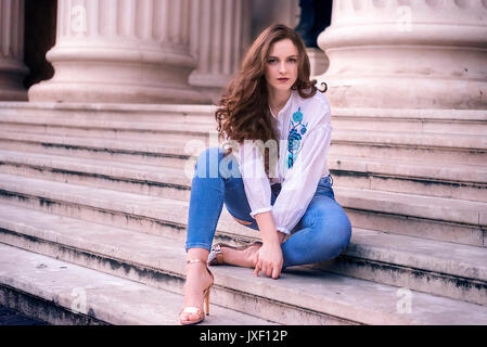 Ragazza giovane con capelli castani e occhi blu, si siede sul vecchio gradini in marmo vestito moderno e guardando la telecamera. Foto Stock