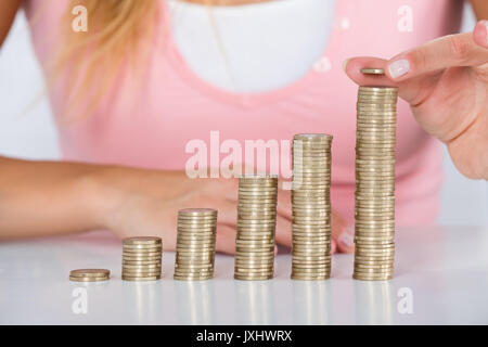 Close-up di donna mano inserire moneta sulla pila di monete a scrivania bianca Foto Stock
