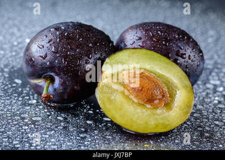 Per due e una metà a fette Damson prugne con la pietra su un nero tagliere con gocce d'acqua. Macro shot di fresco, organica frutta estiva. Foto Stock