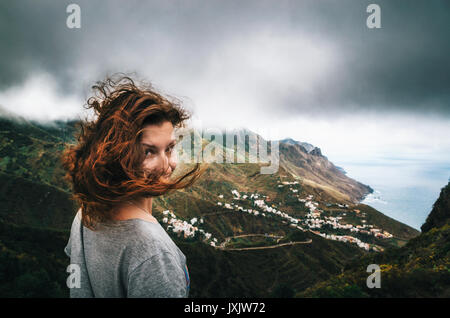 Gioiosa giovane donna con capelli volanti nascosto il suo volto guarda la telecamera di fronte Taganana villaggio sul nord di Tenerife. Nozione di viaggio, happ Foto Stock