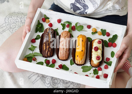 Donna che mantiene in legno bianco vassoio pieno di francese eclairs seduto sul divano Foto Stock