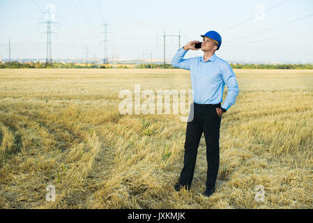 Da parte di un tecnico o di un lavoratore sorrisi nel casco protettivo in possesso di un telefono cellulare. Campi di grano, linea di alimentazione, giornata di sole e cielo blu. Foto Stock