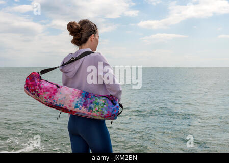 Vista posteriore di una donna che porta un materassino yoga sacchetto, che guarda al mare. Foto Stock