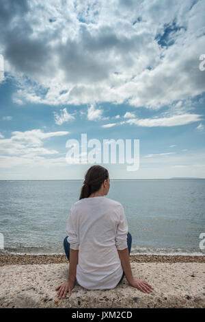 Vista posteriore di una donna seduta su una parete che guarda al mare Foto Stock