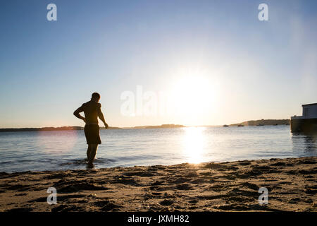 Retroilluminato maschio giovane nuotatore passeggiate in mare al tramonto Foto Stock