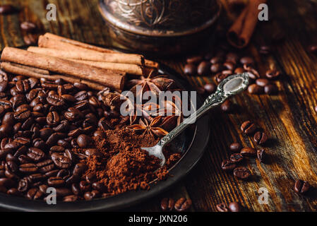 I chicchi di caffè con Spooonful di caffè macinato, bastoncini di cannella e il cinese di anice stellato su piastra in metallo. Alcuni grani sparsi sul tavolo in legno e Cezve su Foto Stock