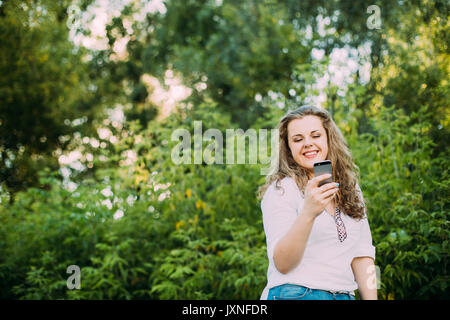 Giovane bella bella Plus dimensioni ragazza caucasica Donna vestita in camicia bianca e jeans blu per godersi la vita, sorridente e utilizza lo smartphone in piedi su S Foto Stock