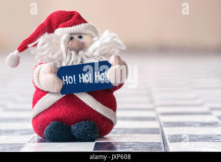 Santa Claus, ripiene fluffy toy, indossando il rosso, con la barba e cappello, tenendo un cartello scritto ho ho ho, sul pavimento piastrellato di nero e bianco, con copia supplementare Foto Stock