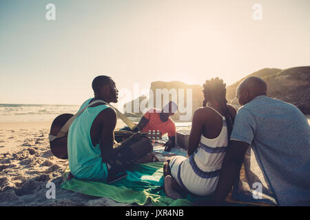 African giovane uomo di cantare e suonare la chitarra sulla spiaggia. Un gruppo di quattro persone aventi una grande volta al picnic sulla spiaggia. Foto Stock