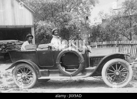 Foto di paesaggio a lunghezza intera di due uomini afroamericani maturi in un'automobile, un uomo nella parte posteriore, un uomo al volante, un bambino caucasico nella parte anteriore, all'aperto, espressioni facciali neutre, 1920. Foto Stock