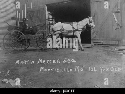 Ritratto a lunghezza intera di un uomo afro-americano in buggy guidato da cavallo, con abiti scuri, seduto accanto alla porta aperta del fienile, espressione neutra, 1915. Foto Stock