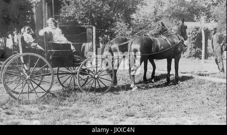 Ritratti a lunghezza intera di giovane afroamericana e giovane bianca, entrambi con abiti leggeri, a cavallo con due cavalli scuri, afroamericano in piedi davanti a cavalli, con vestito e cappello scuri, espressioni neutre, 1920. Foto Stock