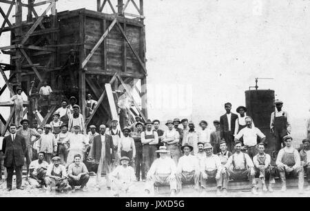 Un folto gruppo di lavoratori americani con espressioni di folle viene raccolto al di fuori di fronte e su una grande torre di legno struttura, 1915. Foto Stock