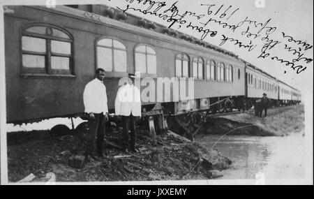 Due americano africano di addestrare i dipendenti in piedi di fronte a un treno Il treno è di attraversare un piccolo ponte con acqua al di sotto di una ferrovia lavoratore è sull'altro lato della patch di acqua, due dipendenti sono luce da indossare magliette colorate e di colore scuro pantaloni, 1912. Foto Stock