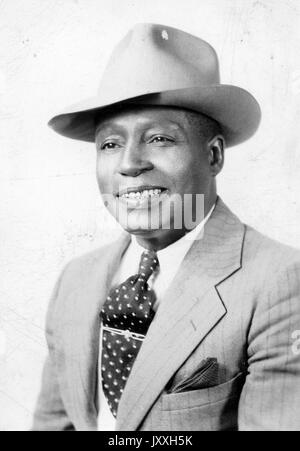 Headshot di uomo afroamericano maturo, con giacca a righe scure, fazzoletto, cravatta scura con design, clip per cravatta, camicia e cappello chiari, espressione sorridente, 1925. Foto Stock