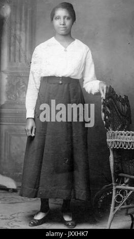 Ritratto di una donna afro-americana che si trova di fronte ad un murale con la mano sinistra che riposa sul retro di una sedia in vimini, il suo braccio destro è al suo fianco, lei indossa una lunga gonna di colore scuro e una blusa a manica lunga di colore chiaro, 1915. Foto Stock