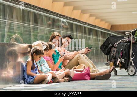 La famiglia in attesa sul piano dell'aeroporto utilizzando i telefoni cellulari Foto Stock