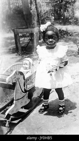 Ritratto di una ragazza afro-americana a lunghezza intera, con un abito leggero, calzini e archetto, fiori in piedi, in piedi all'aperto accanto a un cavaliere a molla che porta la bambola, espressione neutra, 1925. Foto Stock