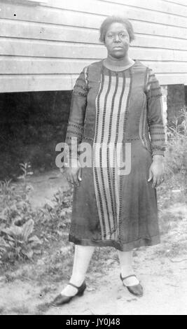 Ritratto di una donna afro-americana in piedi di fronte ad una struttura elevata, è vestita piacevolmente, indossando un lungo abito, collant, e scarpe da vestito a chioccarpa di colore scuro, 1915. Foto Stock