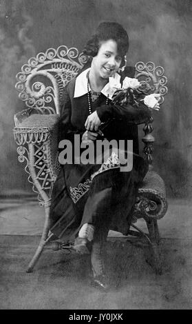 Ritratto di una giovane donna afroamericana seduta in una sedia di vimini, le sue gambe sono incrociate e sta tenendo un mazzo di fiori nel braccio sinistro, lei indossa un vestito di colore scuro con un grande collare di colore chiaro, lei è sorridente, 1915. Foto Stock