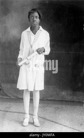 Ritratto a lunghezza intera di una giovane afroamericana, con un abito bianco e calze bianche, in piedi davanti a uno sfondo, con un diploma arrotolato in mano, con una grave espressione facciale, 1929. Foto Stock