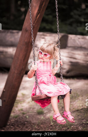 Sweet little girl, vestito con un abito rosa, gode lo swing su un parco giochi in una giornata di sole. La foto è in colori pastello. Foto Stock