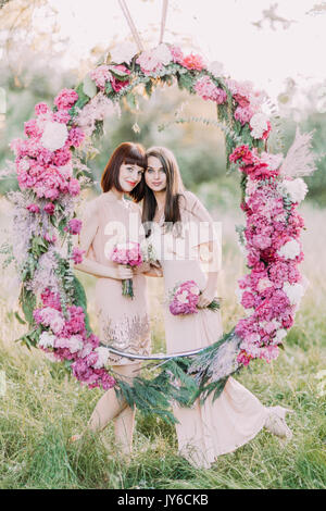 Il bel ritratto di due damigelle tenendo i mazzi di fiori di colore rosa e in piedi dietro il matrimonio di peonie arch collocato nel soleggiato parco Foto Stock