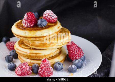 Pila di pancake con lamponi e mirtilli rivestite con zucchero a velo sulla piastra bianca e sfondo nero Foto Stock