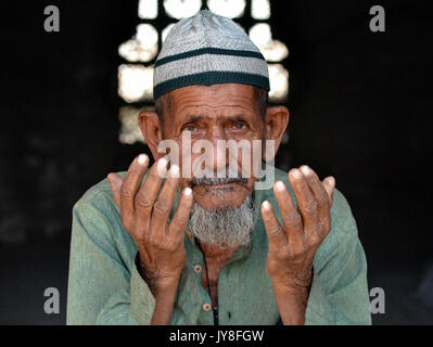 Il vecchio musulmano indiano con la faccia vissuta indossa una capsula di preghiera islamica (taqiyah) e prega con entrambe le mani. Foto Stock