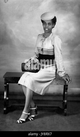 Giovane africano donna americana in un abbigliamento elegante seduta su una panchina e posare per un ritratto in studio con un cappello e una borsa, Kentucky, 1920. Foto Stock