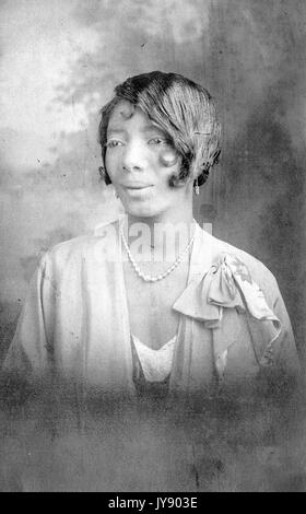 Colpo di testa di matura african donna americana, indossa una camicetta di luce con perla collana e orecchini, espressione neutra, 1925. Foto Stock