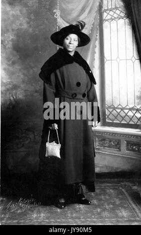 Ritratto di un americano africano woman standing, indossando un grigio cappotto invernale e un cappello nero, portante un portamonete bianco, 1915. Foto Stock