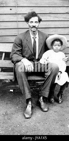 Ritratto di un uomo afro-americano più anziano seduto su una panchina con un bambino, indossa un vestito di colore scuro e un braccio è intorno alla giovane ragazza che indossa un abito di colore chiaro e un cappello, 1920. Foto Stock