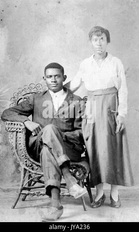 Ritratto di una coppia afro-americana, l'uomo indossa un vestito di colore scuro e si trova su una sedia in vimini con le gambe incrociate, la donna indossa una camicia e una gonna di colore chiaro e si trova accanto a lui con il braccio dietro la schiena, 1920. Foto Stock