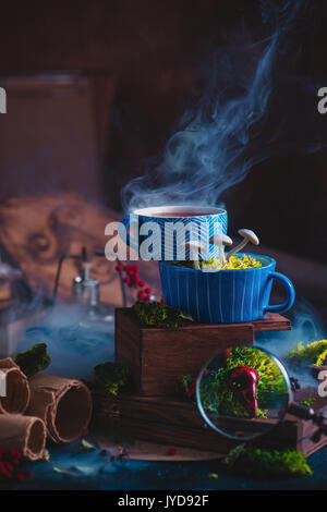 Tazze di ceramica di tè con i funghi in una procedura guidata sul luogo di lavoro. magic ancora vita con fumo, cartigli bird teschi e lente di ingrandimento. Foto Stock