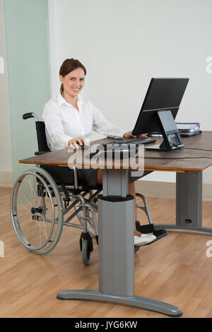 Felice imprenditrice seduto su una sedia a rotelle mentre si lavora sul computer alla reception Foto Stock