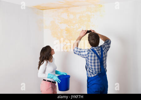 Tuttofare con la donna fotografare acqua danneggiato il soffitto con telefono cellulare a casa Foto Stock