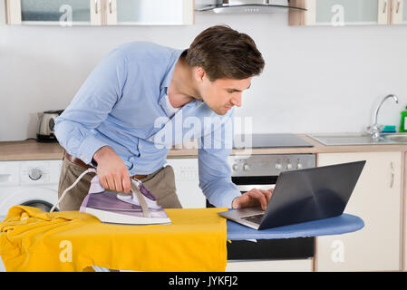 Giovane uomo digitando su laptop durante la stiratura panno in cucina Foto Stock