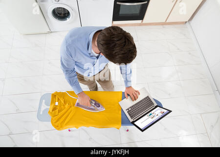 Giovane uomo chat sul sito web sociale con il computer portatile durante la stiratura T-shirt Foto Stock